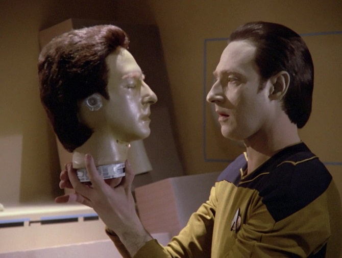 Data from Star Trek holding a robot head and looking at it with curiosity.