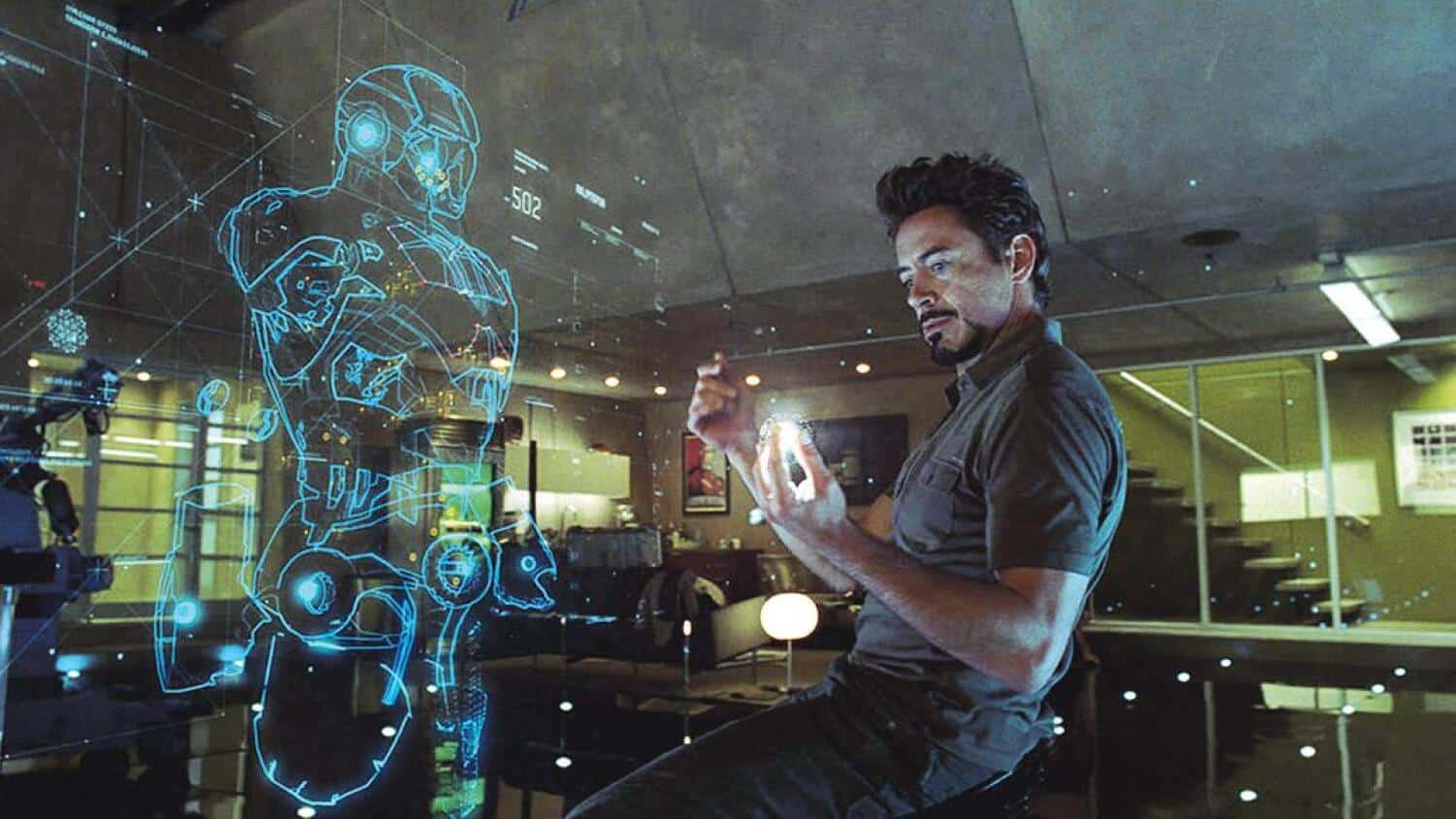 Robert Downey Jr. as Tony Stark in Iron Man, designing a suit of flying armor with the help of his digital assistant JARVIS. A holographic image of a wireframe suit is beside Tony as he speaks to Jarvis through a ball of light.